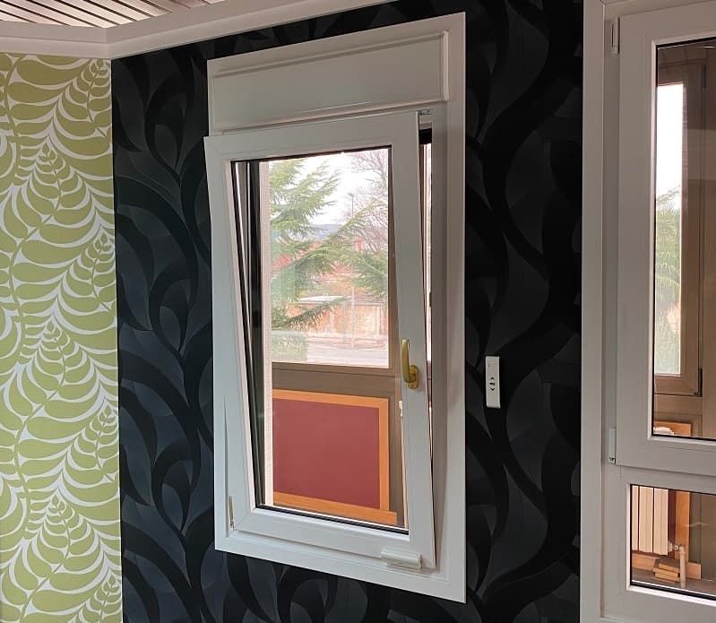 Ventana oscilobatiente con marco blanco, abierta, sobre pared con papel pintado estampado en negro y verde; vista al exterior.