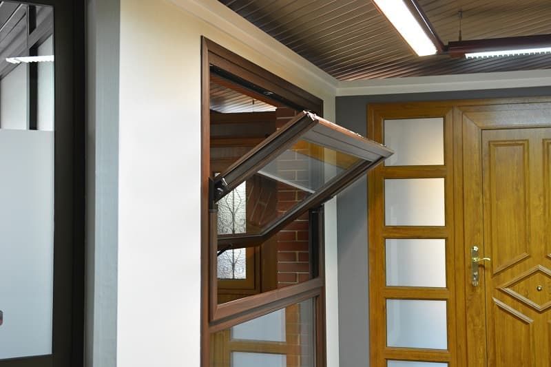 Ventana con marco marrón abierta, revelando una pared de ladrillos, junto a una puerta de madera y una pared blanca.