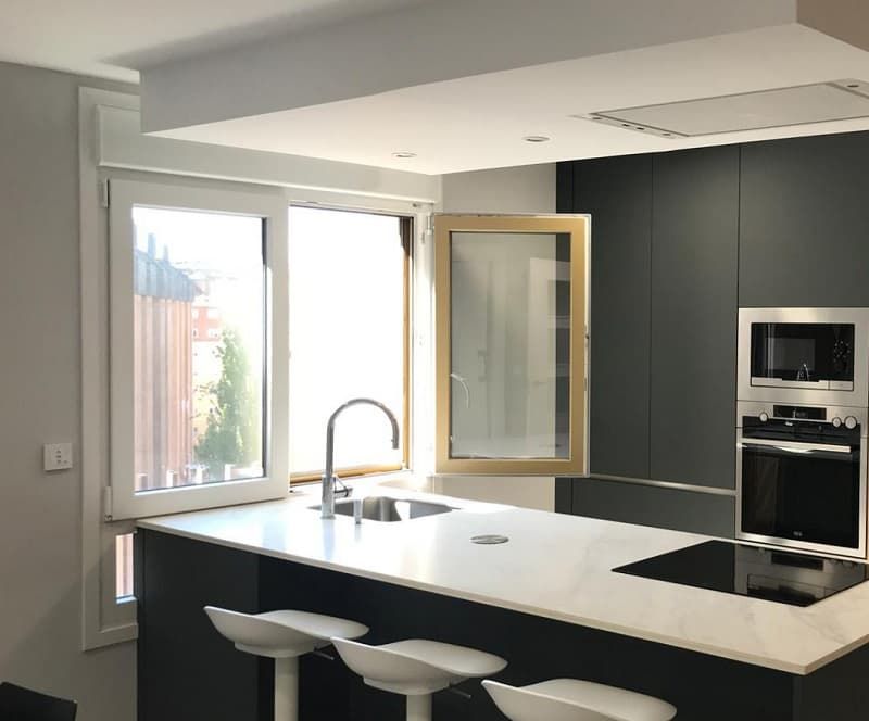 Cocina moderna con ventana abierta sobre el fregadero, gabinetes oscuros, encimera blanca y taburetes de bar.