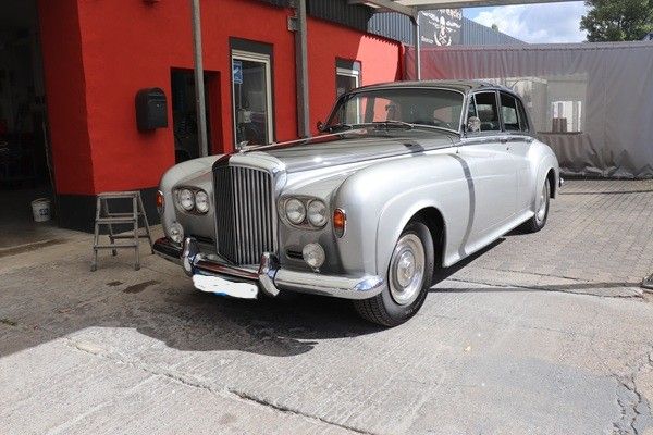 Ein silberner Oldtimer-Rolls-Royce parkt neben einem roten Gebäude im Freien.