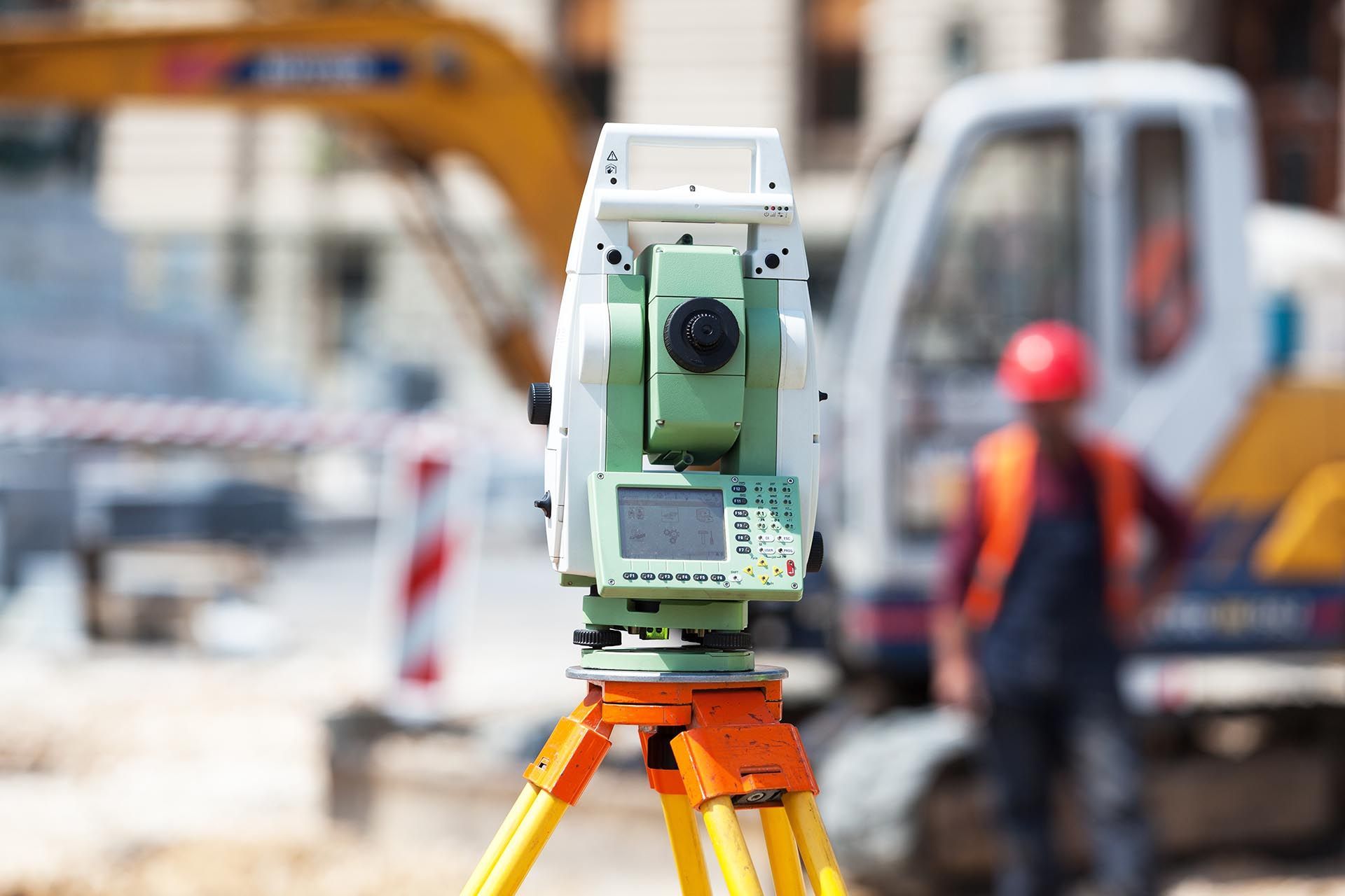 Vermessungsinstrument auf Stativ auf einer Baustelle, im Hintergrund verschwommener Arbeiter und Maschinen.