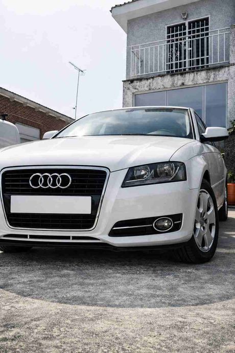 Sedán Audi blanco estacionado frente a una casa con balcón.