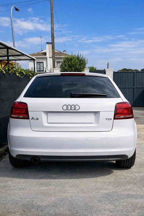 Vista trasera de un sedán Audi blanco estacionado en una entrada de vehículos bajo un cielo azul.