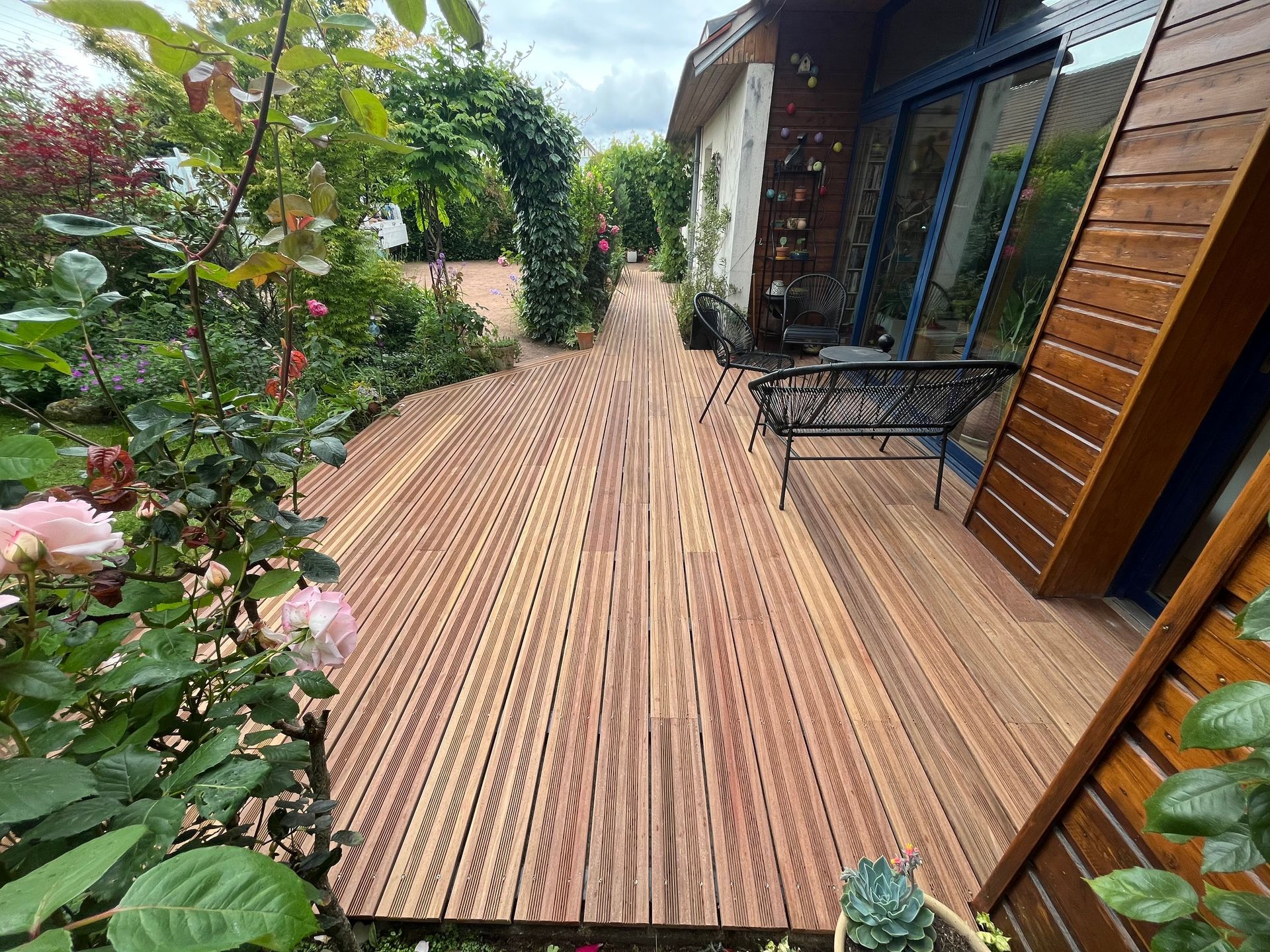 Grande terrasse en bois avec salon noir, maison sur la droite et jardin sur la gauche.