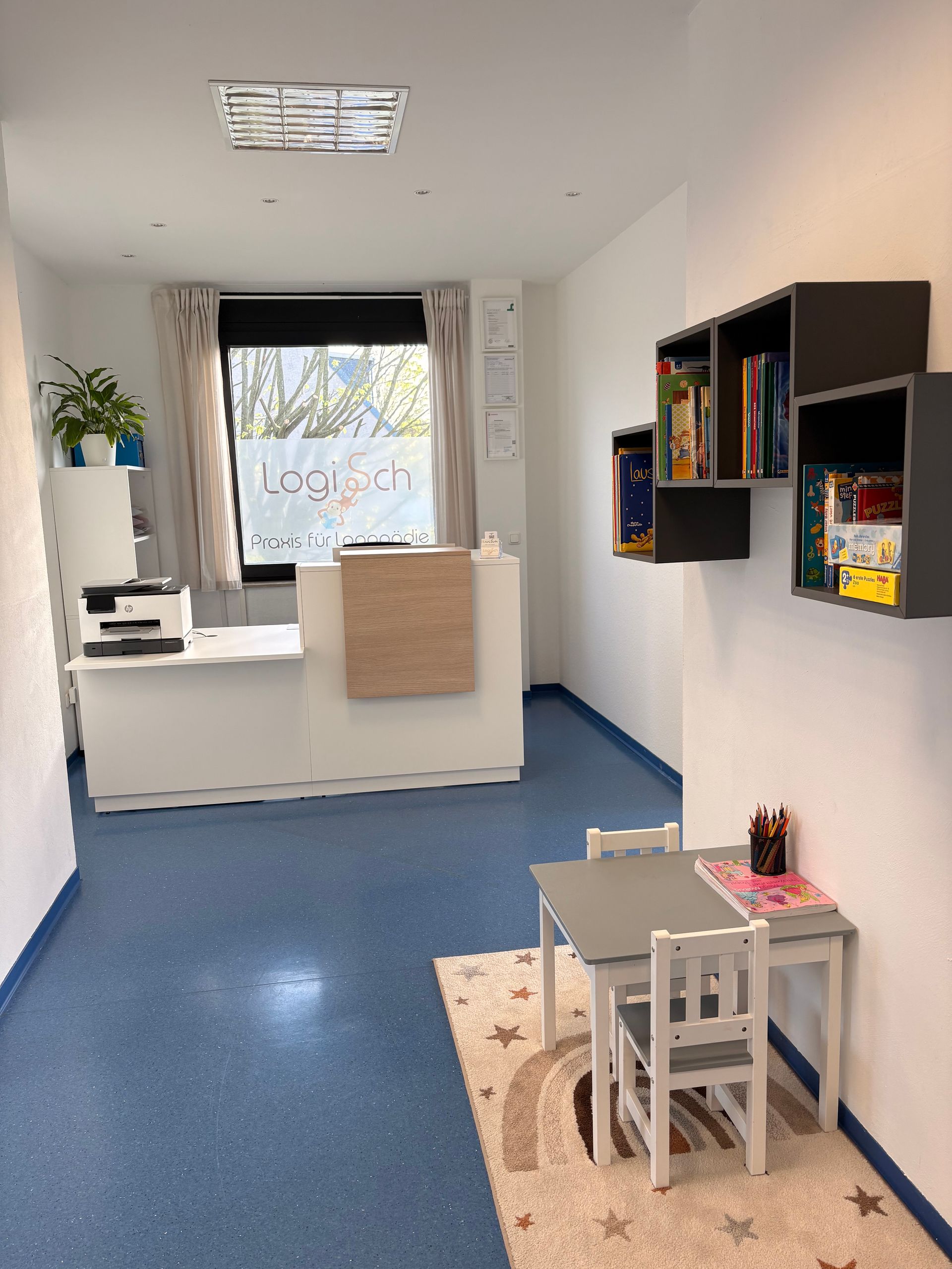 Ein heller Empfangsbereich im Büro mit weißem Schreibtisch, blauem Boden und einem kleinen Kindertisch mit Stuhl auf einem sternengemusterten Teppich.