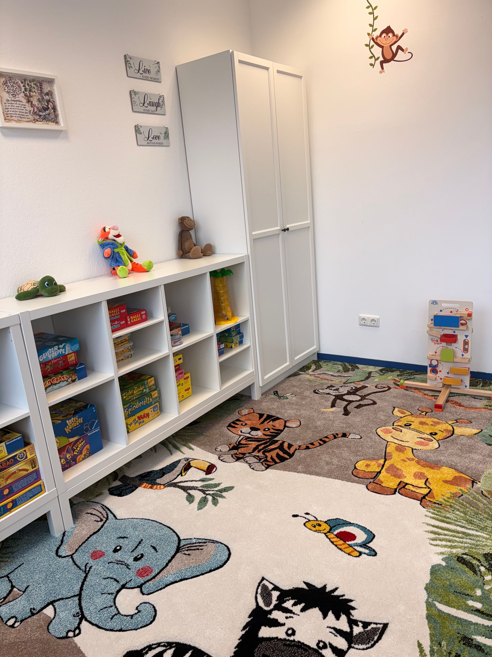 Ein Kinderzimmer mit einem Teppich mit Tiermotiven, einem weißen Spielzeugregal, einem hohen Kleiderschrank und Wandaufklebern mit Tiermotiven.