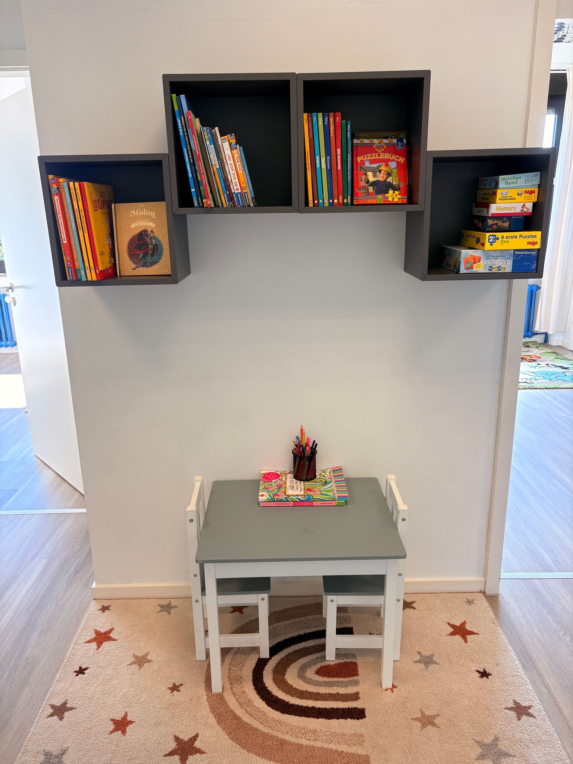 Kleiner Spielbereich mit an der Wand befestigten Spielzeugregalen und einem kindgerechten Tisch mit Stühlen auf einem Teppich.
