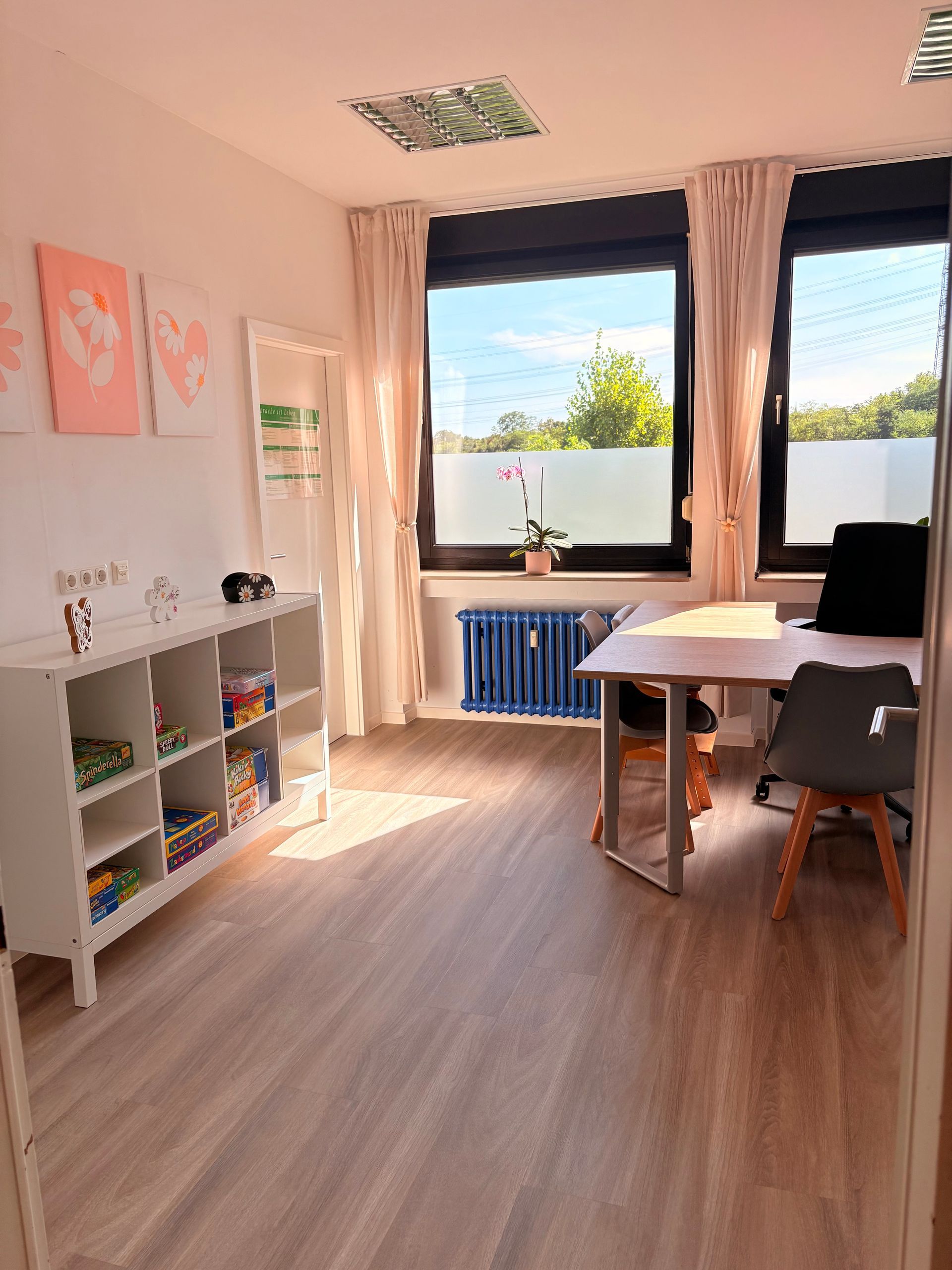 Helles Kinderzimmer mit Holzboden, Schreibtisch an großen Fenstern, Spielzeug und pastellfarbenen Wandbildern.
