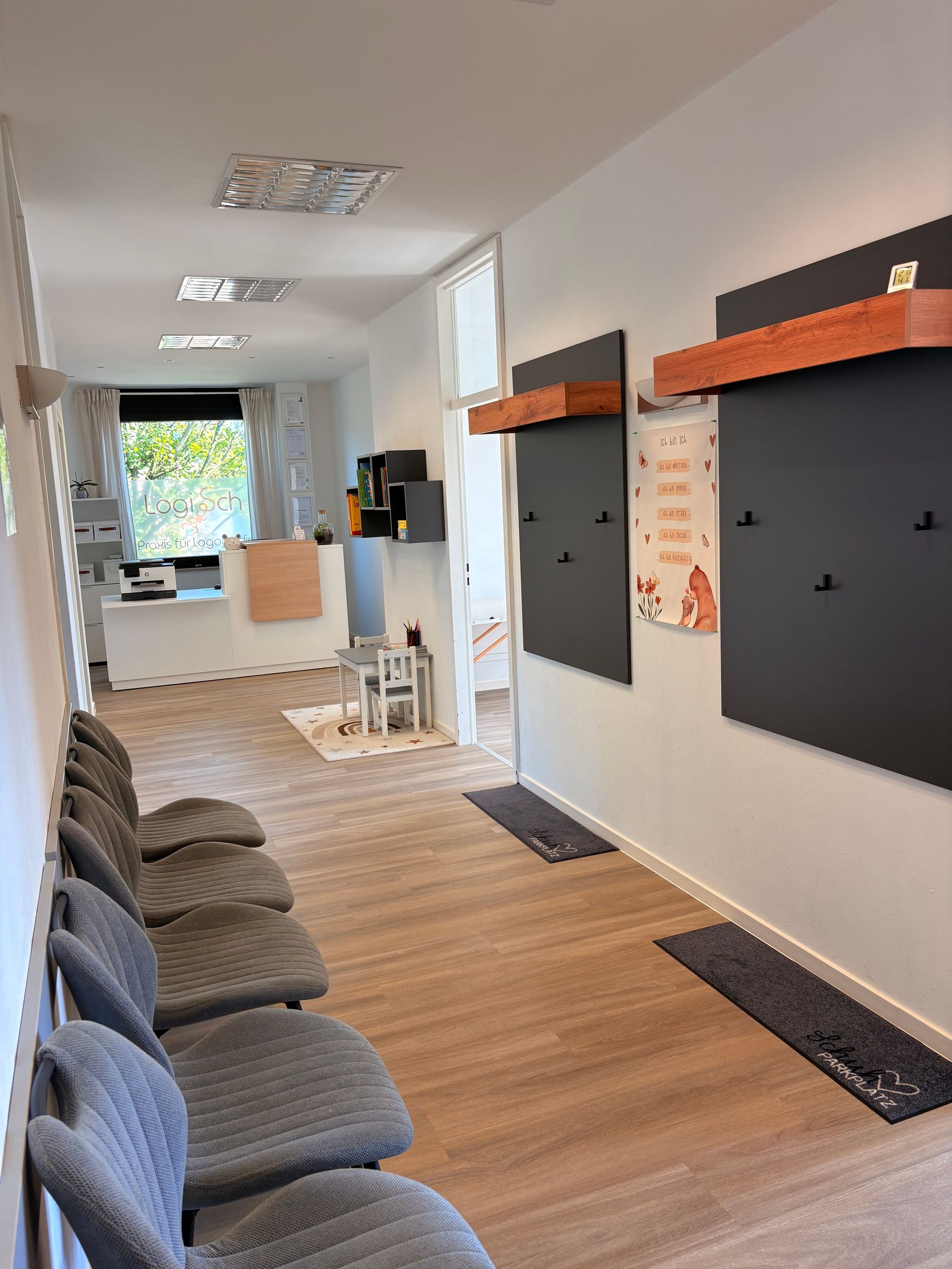 Schmaler Salonflur mit Wandregalen, grauen Stühlen und einem kleinen Hocker in der Nähe eines hellen Fensters.