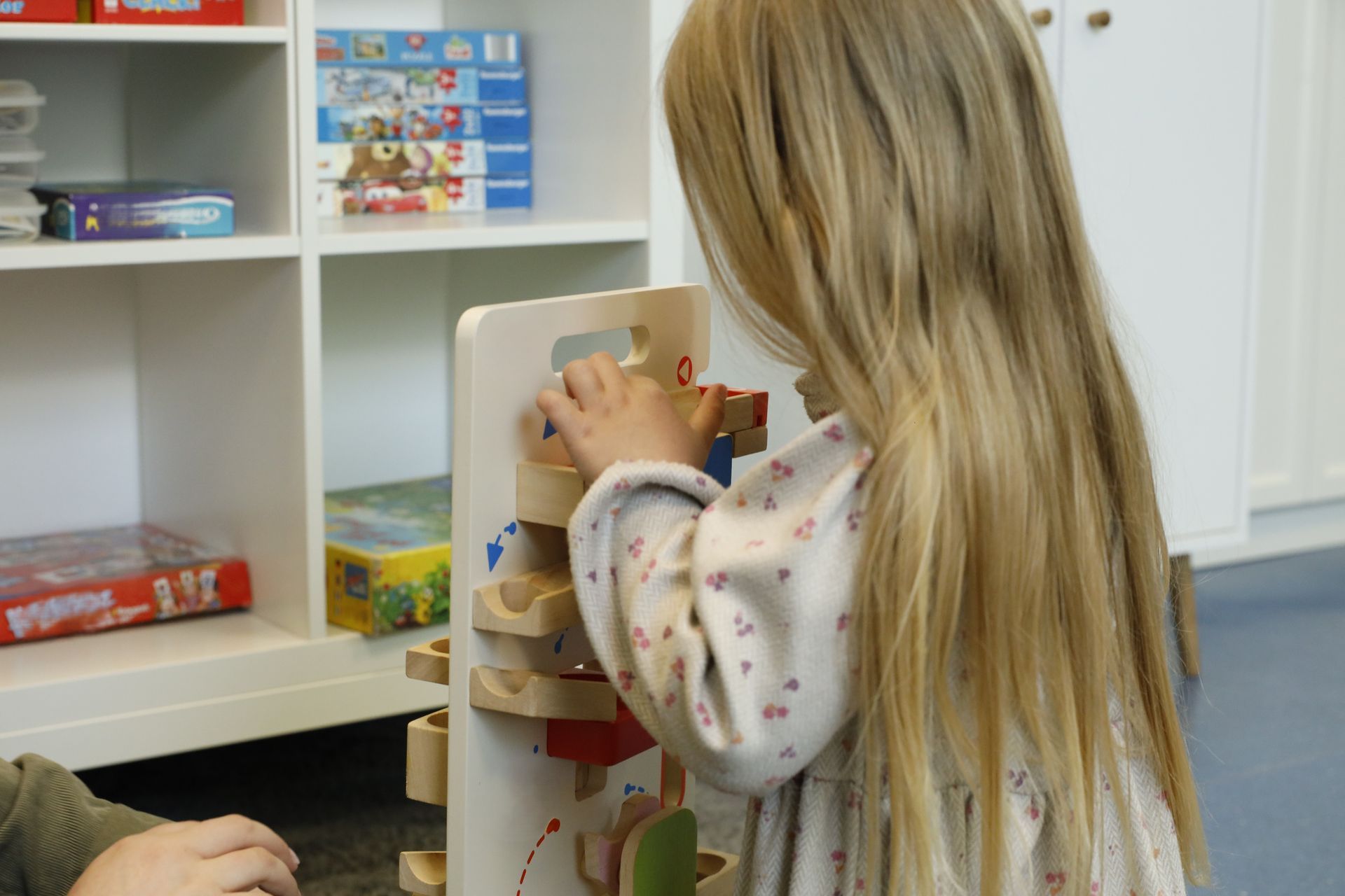 Ein junges Mädchen mit blonden Haaren spielt mit einem Holzspielzeug in einem Raum mit Regalen voller Spielzeug.