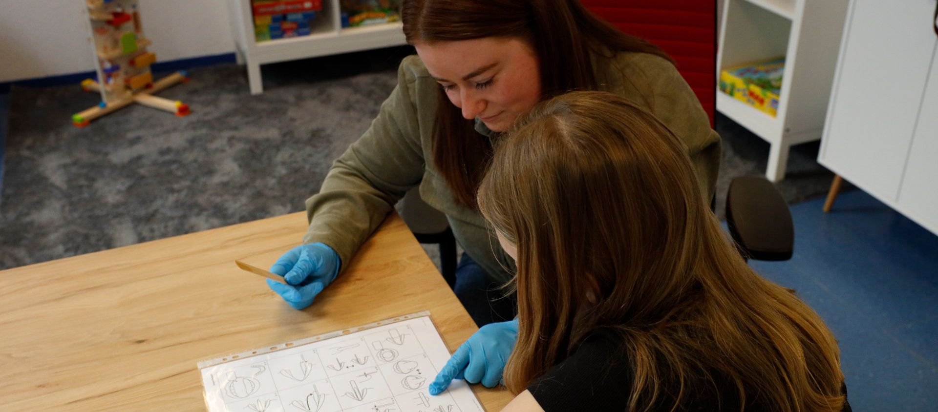 Frau in grünem Hemd und blauen Handschuhen zeigt auf ein Blatt Papier; ein Kind sitzt am Tisch. Heller Raum, Holztisch.
