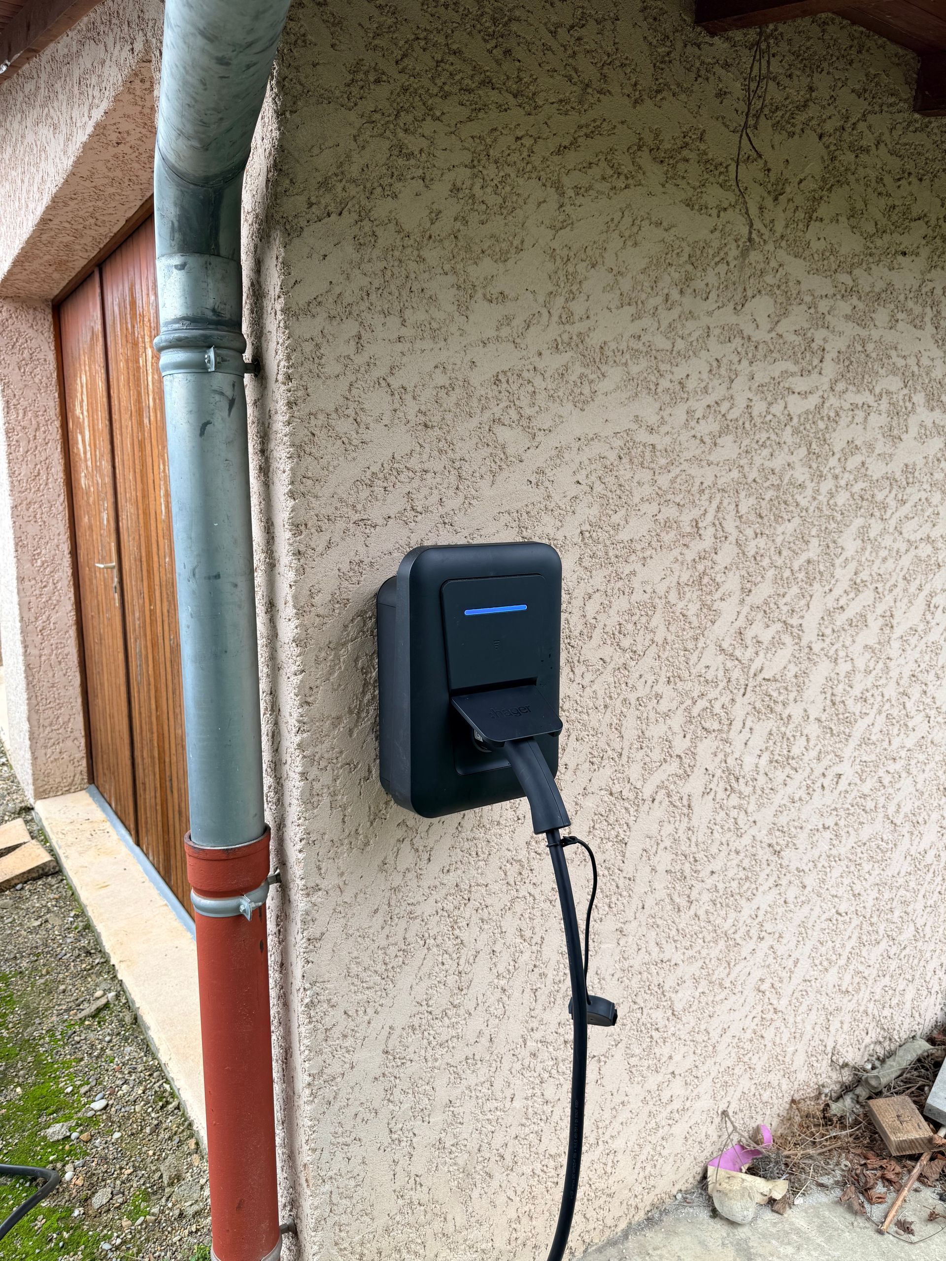Borne de recharge noire pour véhicules électriques, fixée sur un mur en stuc à côté d'une gouttière.