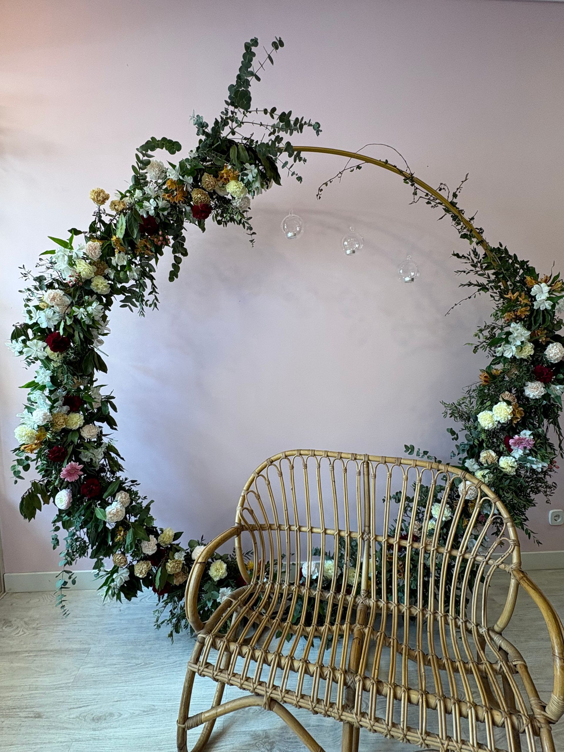 Un banco tejido se encuentra delante de un gran arco circular decorado con flores, frente a una pared de color rosa pálido.