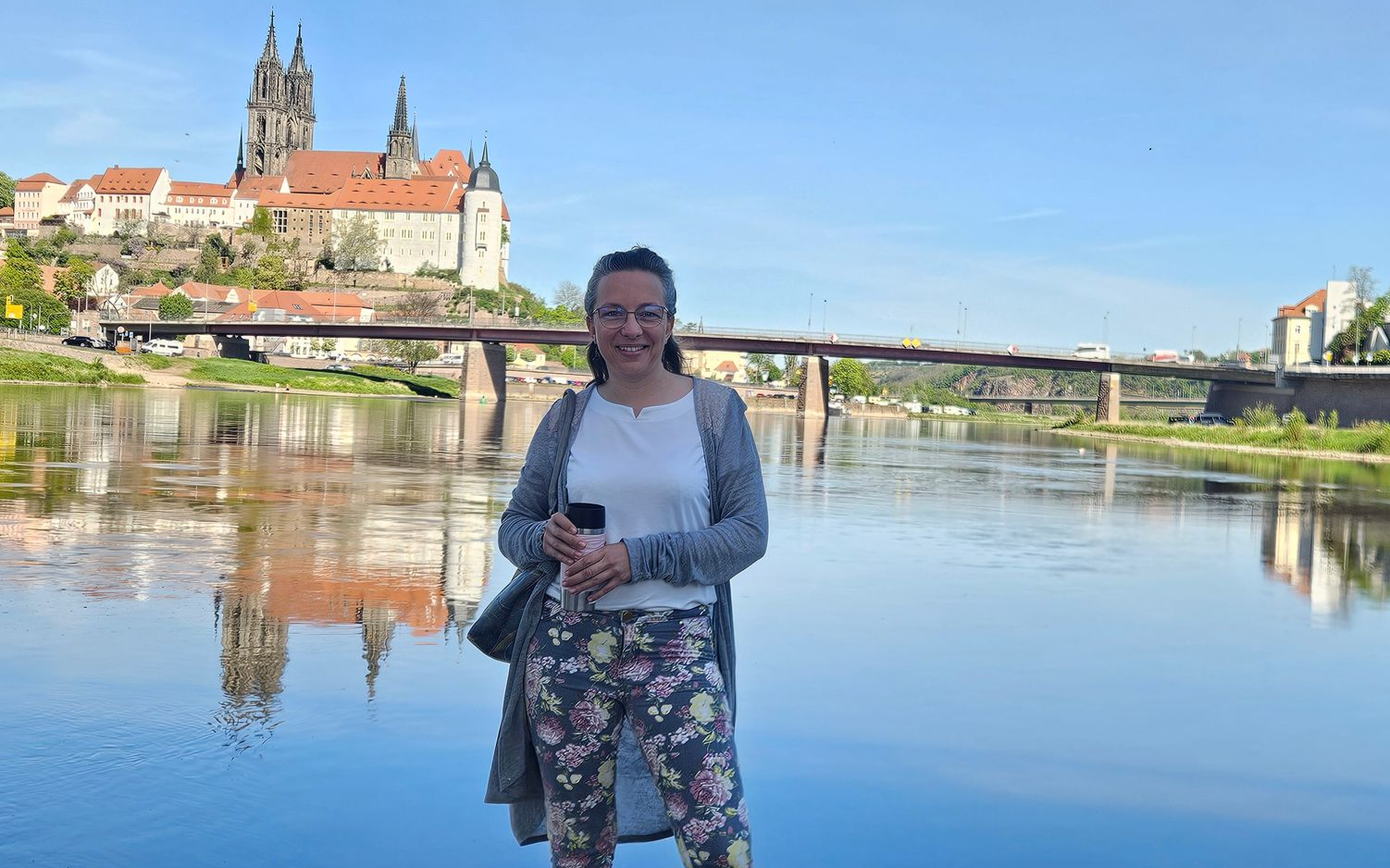 Eine Person steht an einem Fluss, in dessen Wasser sich eine Kathedrale und eine Brücke spiegeln, und hält eine Kamera in der Hand.
