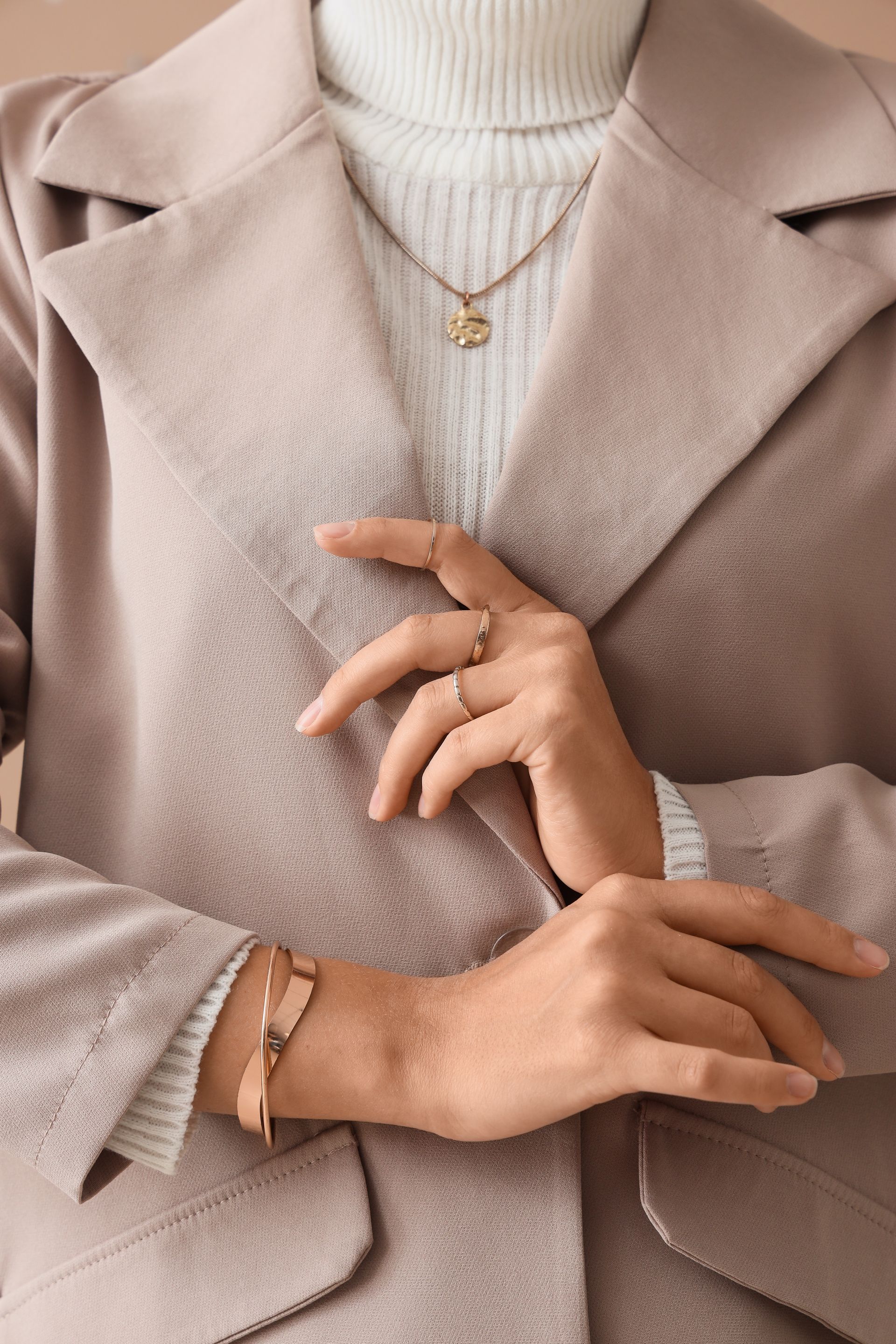 Femme portant une veste beige, un col roulé blanc, des bijoux en or et des bagues, les mains croisées.