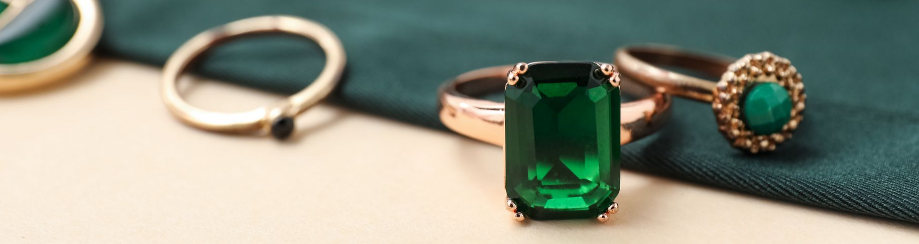 Gros plan de trois bagues en or avec pierres précieuses vertes, reposant sur un tissu vert texturé.