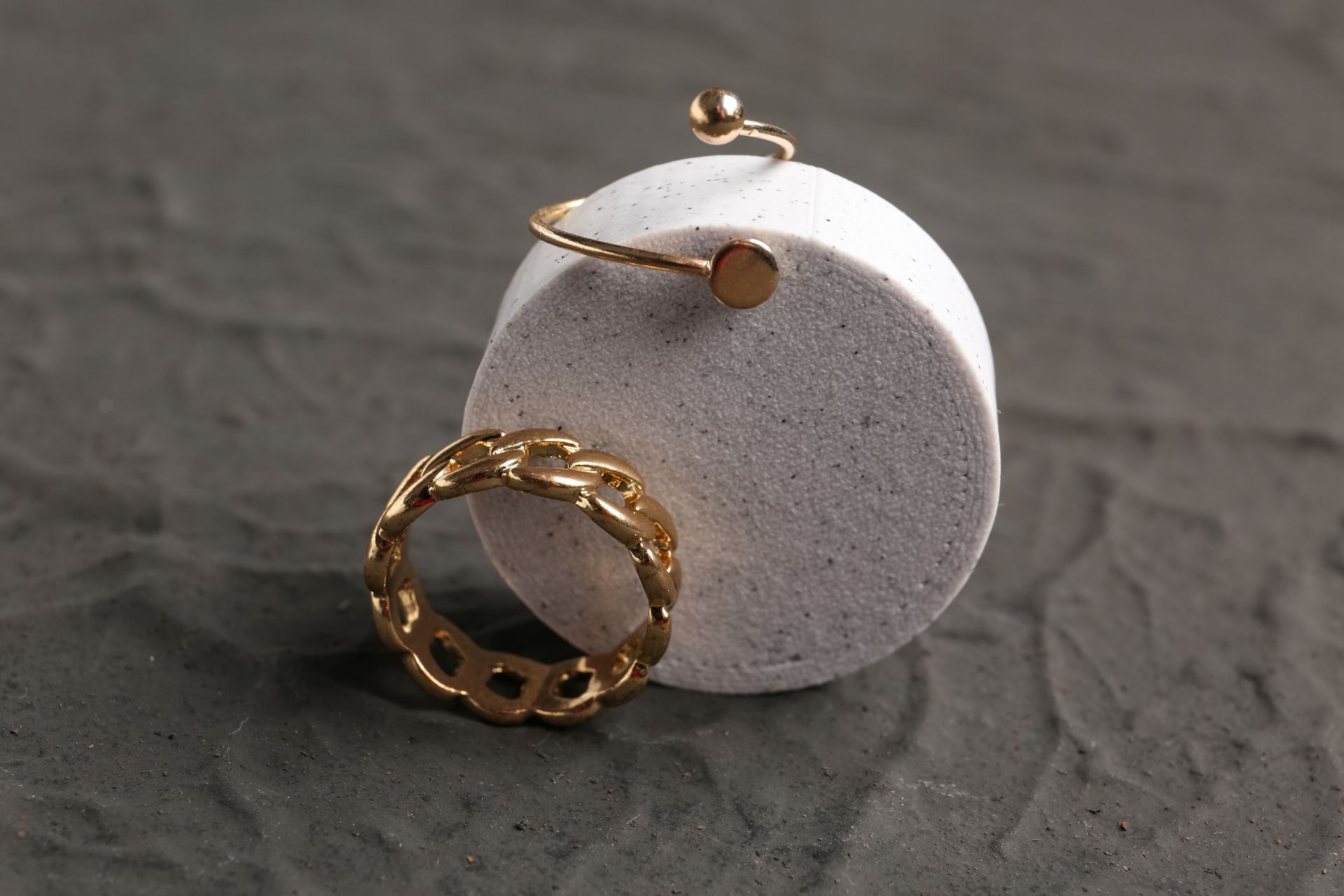 Bijoux en or, bague et bracelet, sur un objet cylindrique blanc sur fond gris texturé.