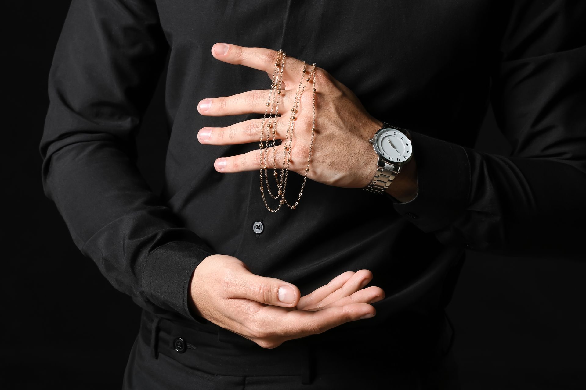 Personne en chemise noire tenant un chapelet en or ; montre au poignet, sur fond noir.