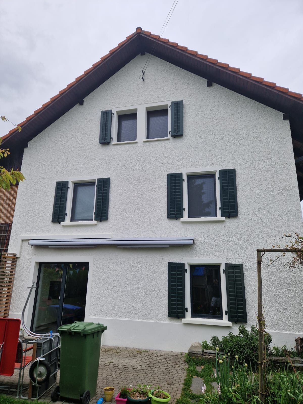 Ein weißes Haus mit schwarzen Fensterläden und einer grünen Mülltonne davor.