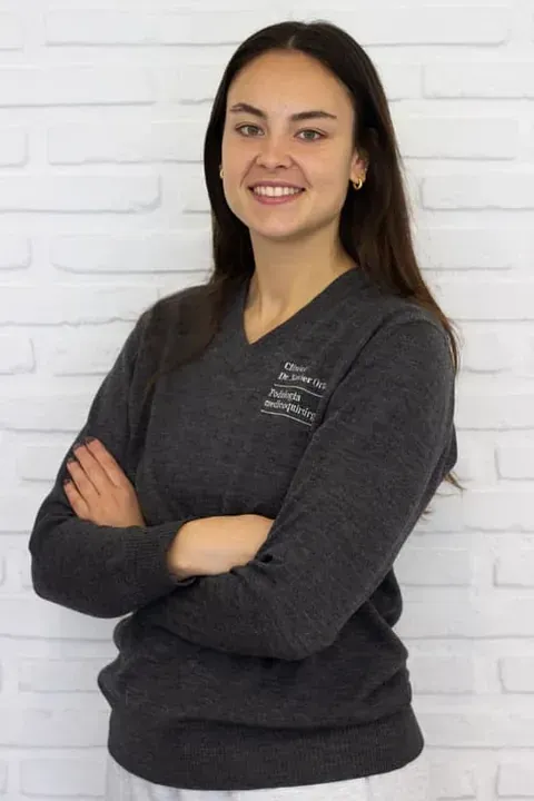 Una mujer con un suéter gris está parada con los brazos cruzados y sonriendo.
