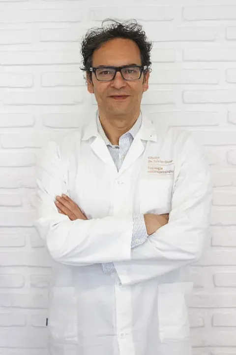 Un hombre con una bata blanca y gafas está de pie con los brazos cruzados.