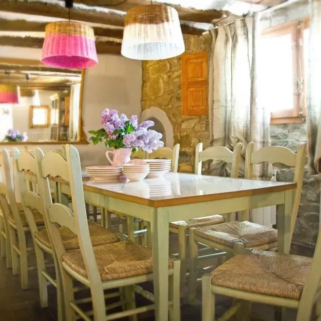 Comedor rústico con mesa larga, sillas, pared de piedra y lámparas rosas y blancas.