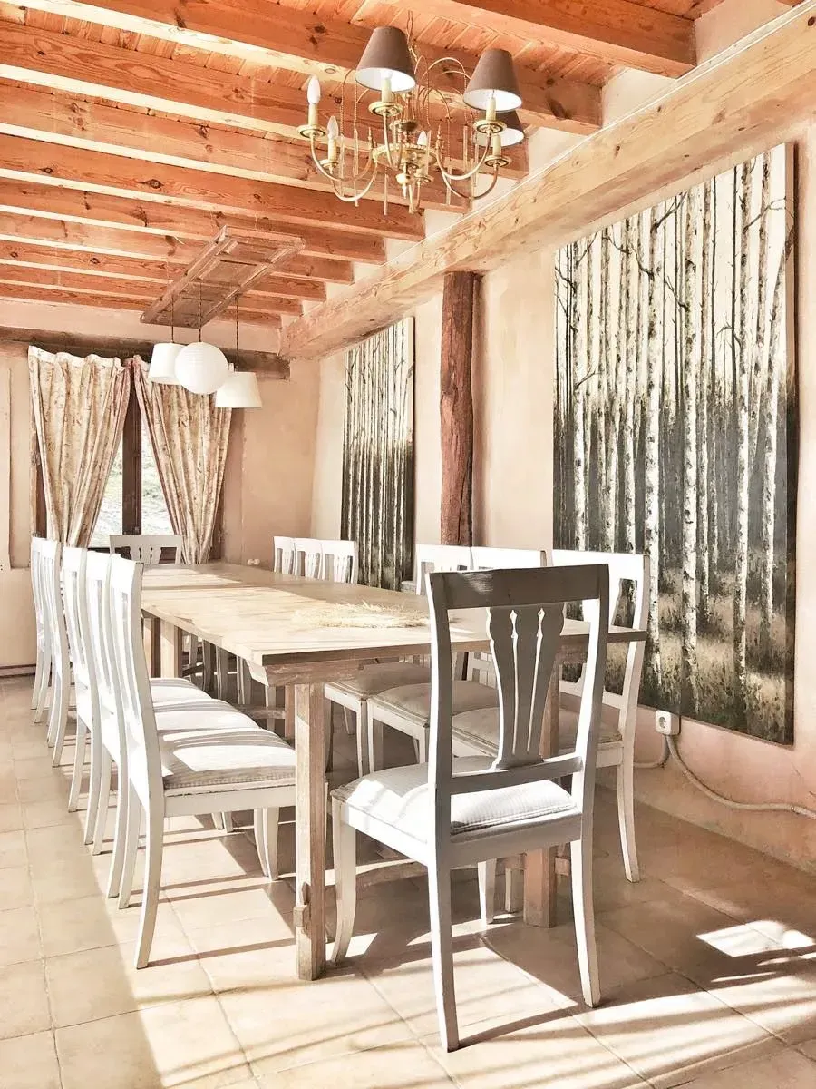 Mesa de comedor larga de madera con sillas blancas en una habitación rústica con vigas de madera, una lámpara de araña y arte de abedul.