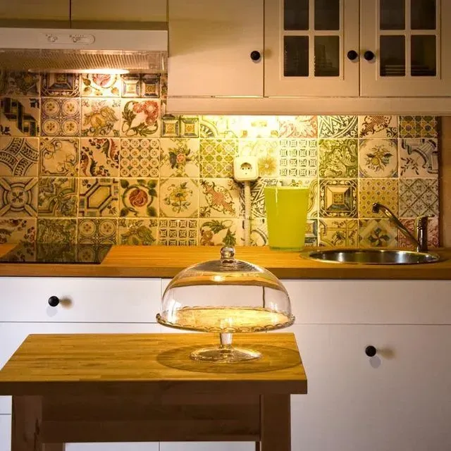 Cocina con salpicadero de azulejos decorativos, gabinetes blancos y una encimera de madera con soporte de vidrio para pastel.
