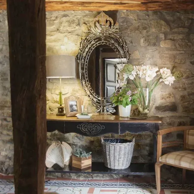Entrada con pared de piedra rústica, consola antigua, espejo ornamentado, lámpara, flores y una silla de madera.