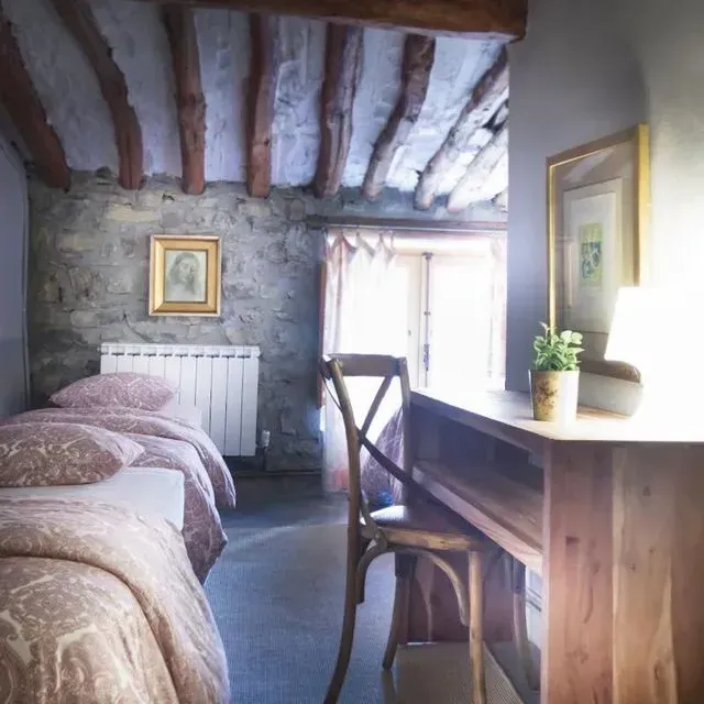 Acogedor dormitorio con paredes de piedra, vigas de madera, dos camas, un escritorio y una ventana.