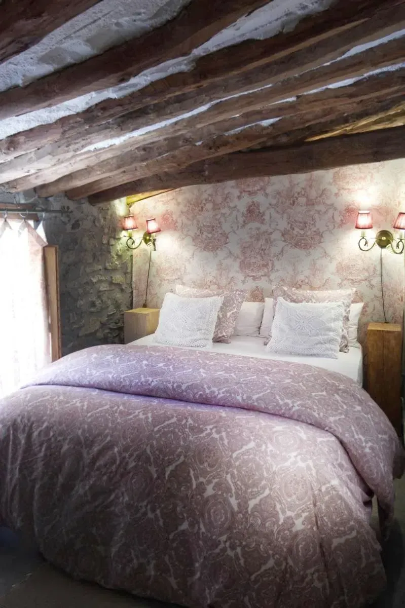 Dormitorio con pared de piedra, vigas de madera, una cama con edredón estampado en color morado y apliques de iluminación.