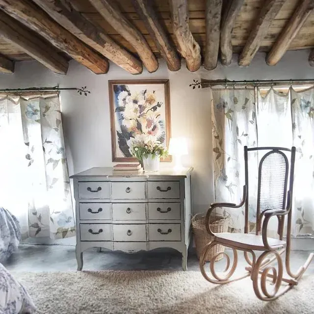 Dormitorio con tocador gris, mecedora, cortinas florales, vigas de madera y un estampado floral enmarcado.