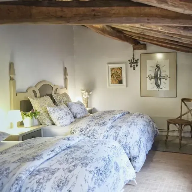 Dormitorio con dos camas, ropa de cama floral azul claro, vigas de madera, arte enmarcado y una lámpara de araña vintage.