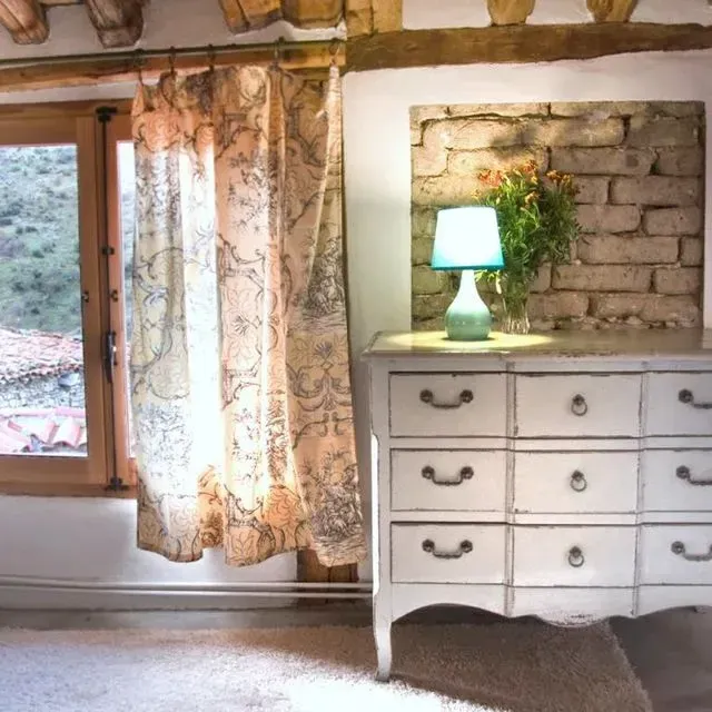 Cómoda blanca con cajones, lámpara turquesa, nicho de ladrillo, ventana con cortina.