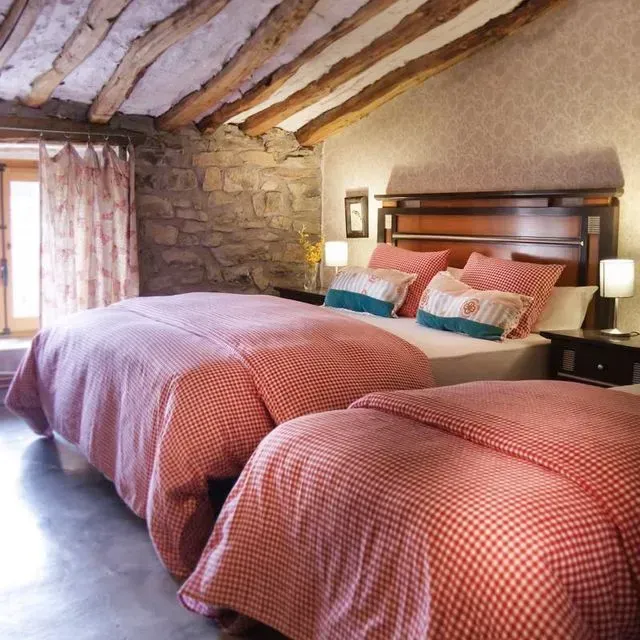 Dormitorio con dos camas, ropa de cama de cuadros rojos, pared de piedra, vigas de madera.