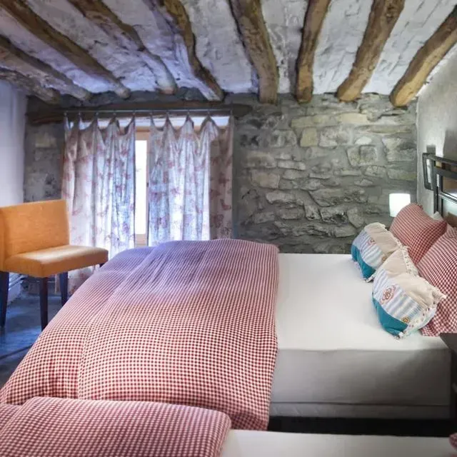 Dormitorio con pared de piedra, vigas de madera, ropa de cama de cuadros rojos y silla naranja.