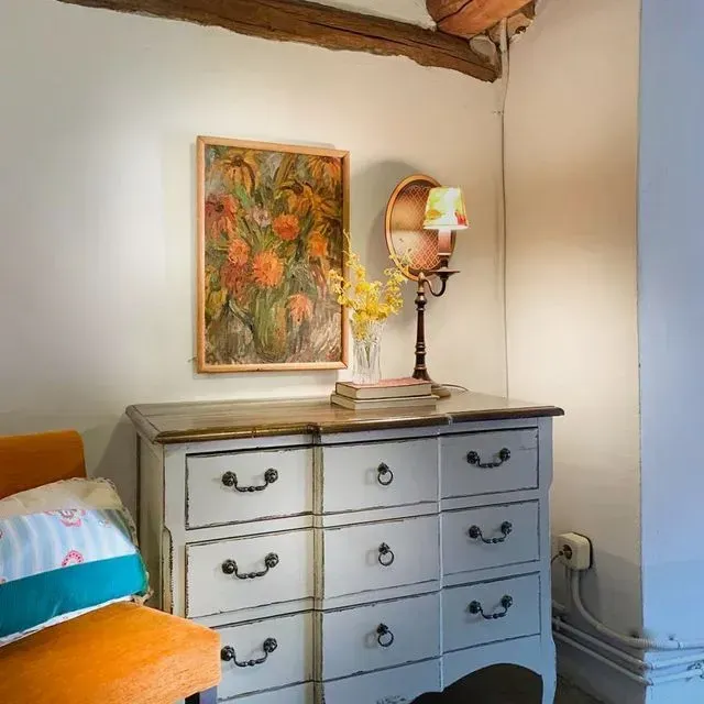 Una cómoda con obras de arte, una lámpara y una silla en un rincón acogedor. Flores y un cojín añaden color.