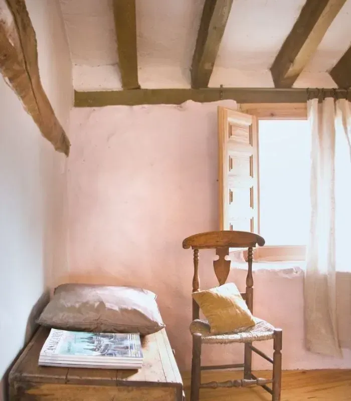 Un dormitorio rústico con paredes rosas, vigas de madera, una silla de madera y un arcón.