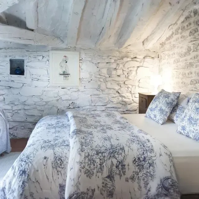 Dormitorio con paredes de piedra blanca, una cama con ropa de cama con motivos florales azules y una obra de arte enmarcada.