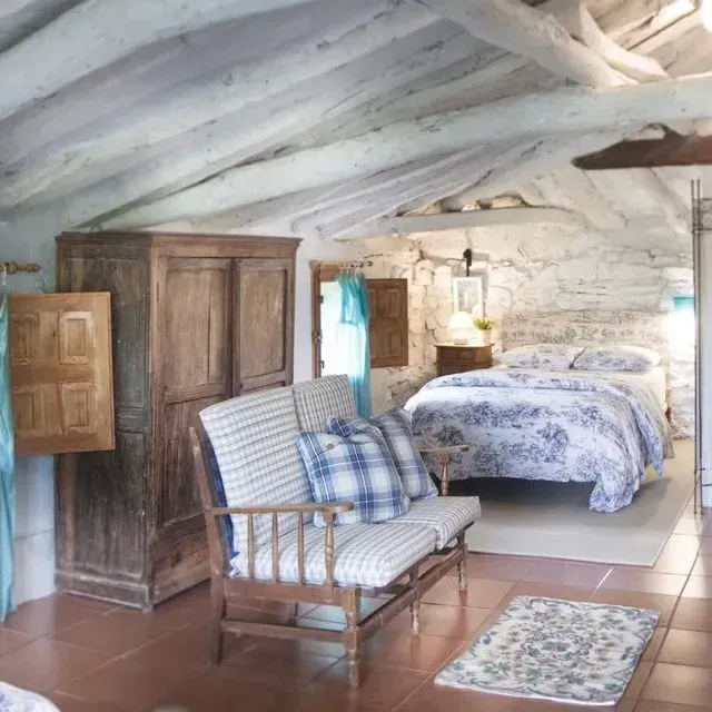 Dormitorio rústico con paredes de piedra, vigas de madera, muebles antiguos y una cama con ropa de cama floral.