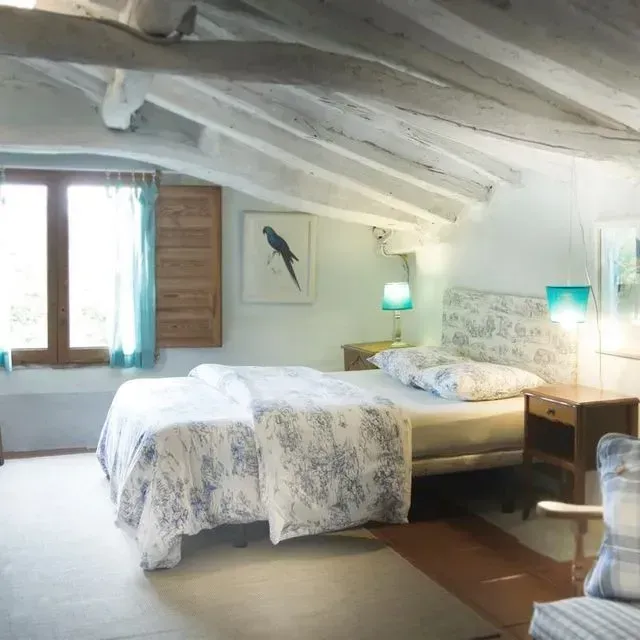 Dormitorio con cama, vigas de madera pintadas de blanco, cortinas azules y obra de arte de un pájaro.