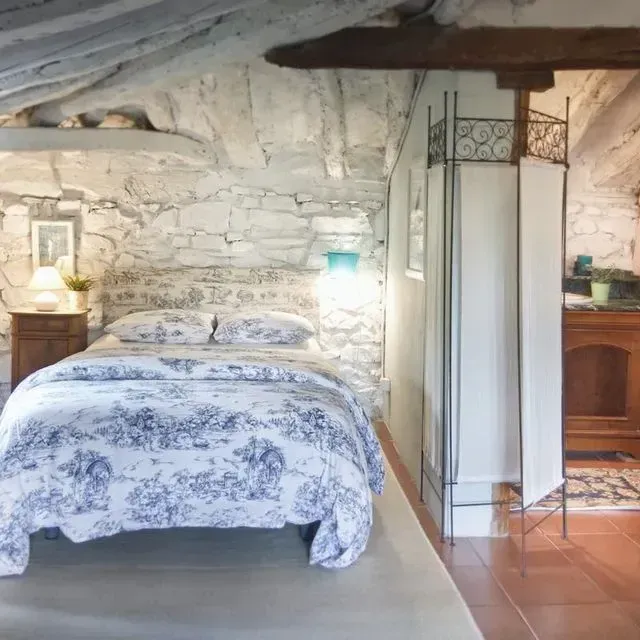 Dormitorio con paredes de piedra, techo de madera, ropa de cama azul y blanca y cortina blanca.