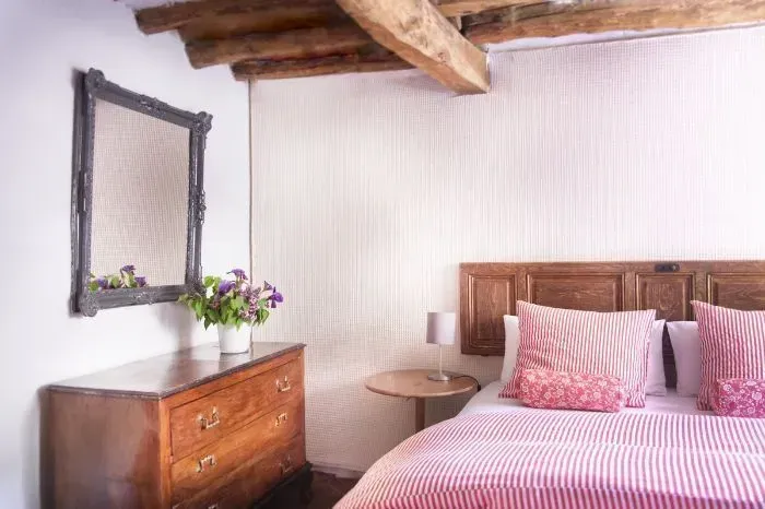 Acogedor dormitorio con vigas de madera, cómoda, cama con ropa de cama de rayas rosas y un espejo.