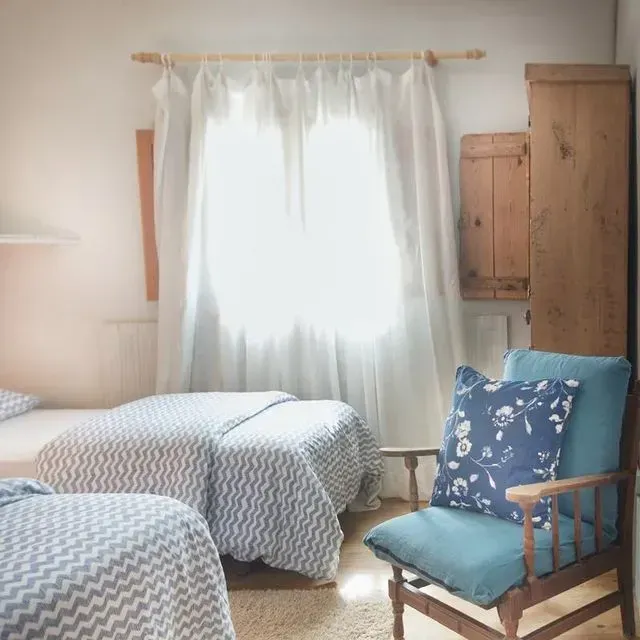 Dos camas individuales con ropa de cama de color azul y una silla azul en un dormitorio luminoso con ventana.