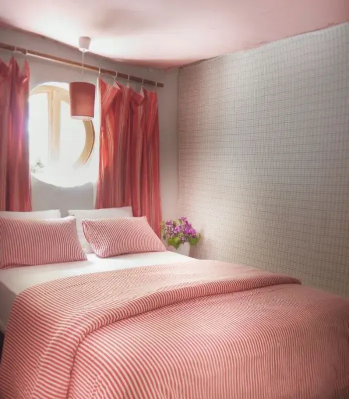 Dormitorio rosa con ropa de cama a rayas, cortinas y una ventana redonda.
