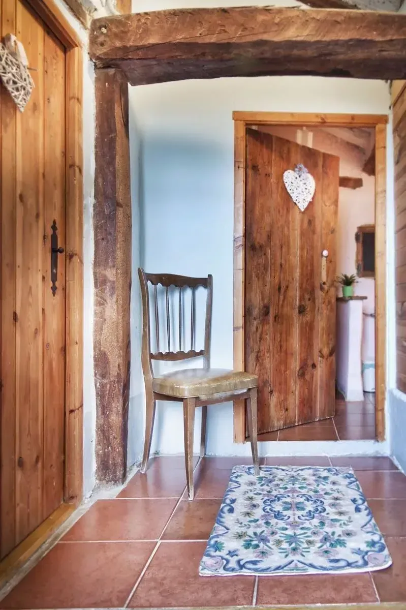 Una entrada rústica con puertas y vigas de madera. Una silla vintage se encuentra junto a una alfombra estampada en azul y blanco.
