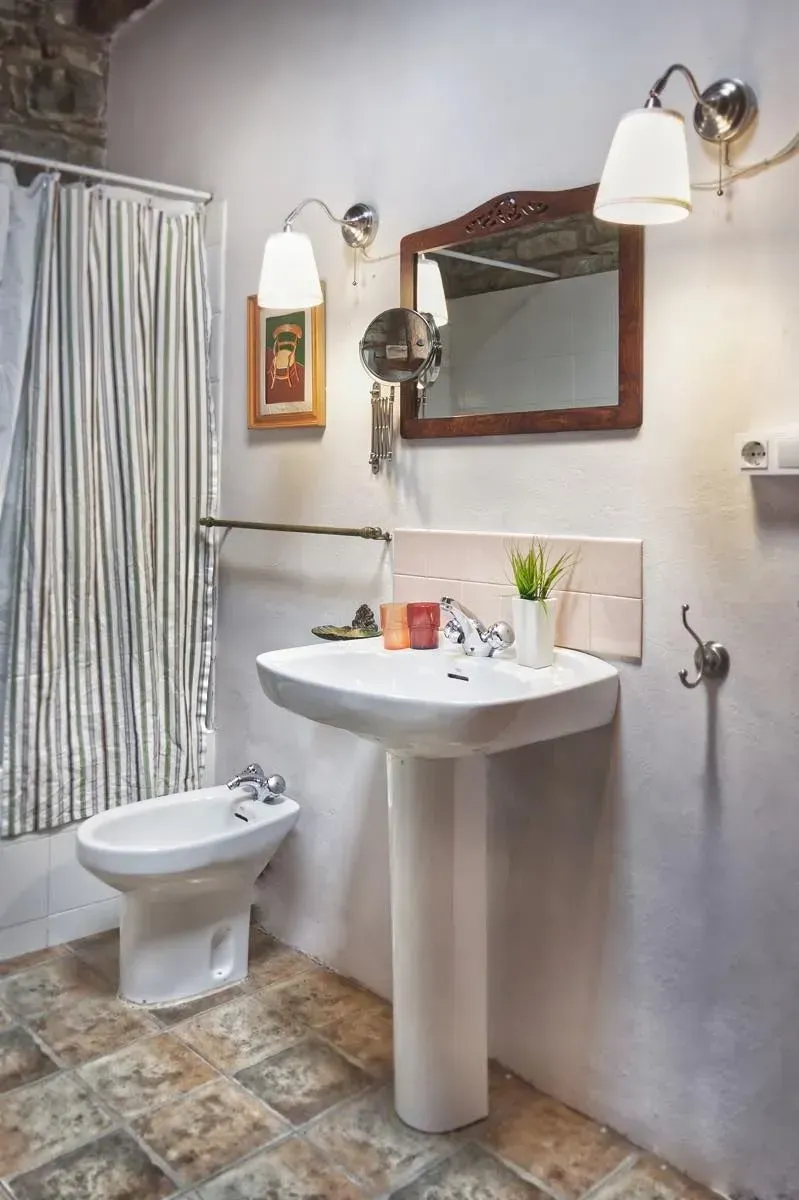 Baño con lavabo de pedestal blanco, bidet, cortina, iluminación de aplique y piso rústico.