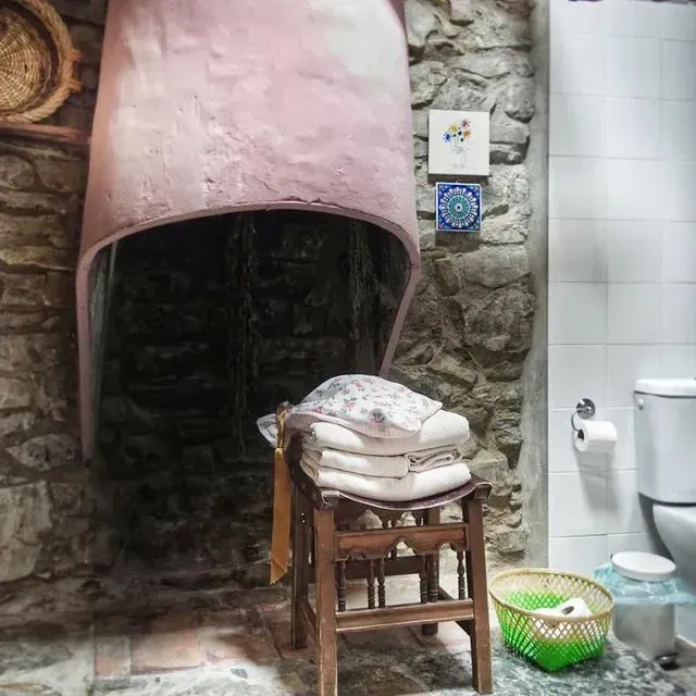 Baño con paredes de piedra, nicho arqueado, taburete de madera con toallas, inodoro y azulejos decorativos.