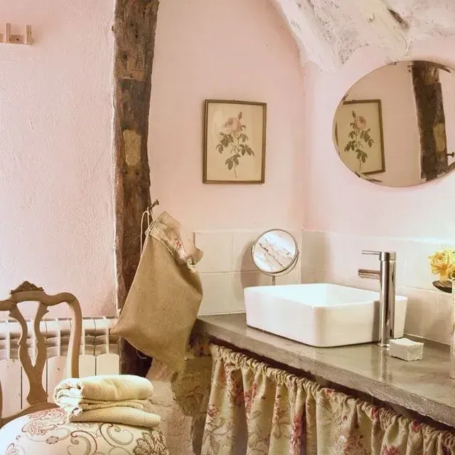 Baño rosa con lavabo blanco, cortinas florales y una silla vintage.