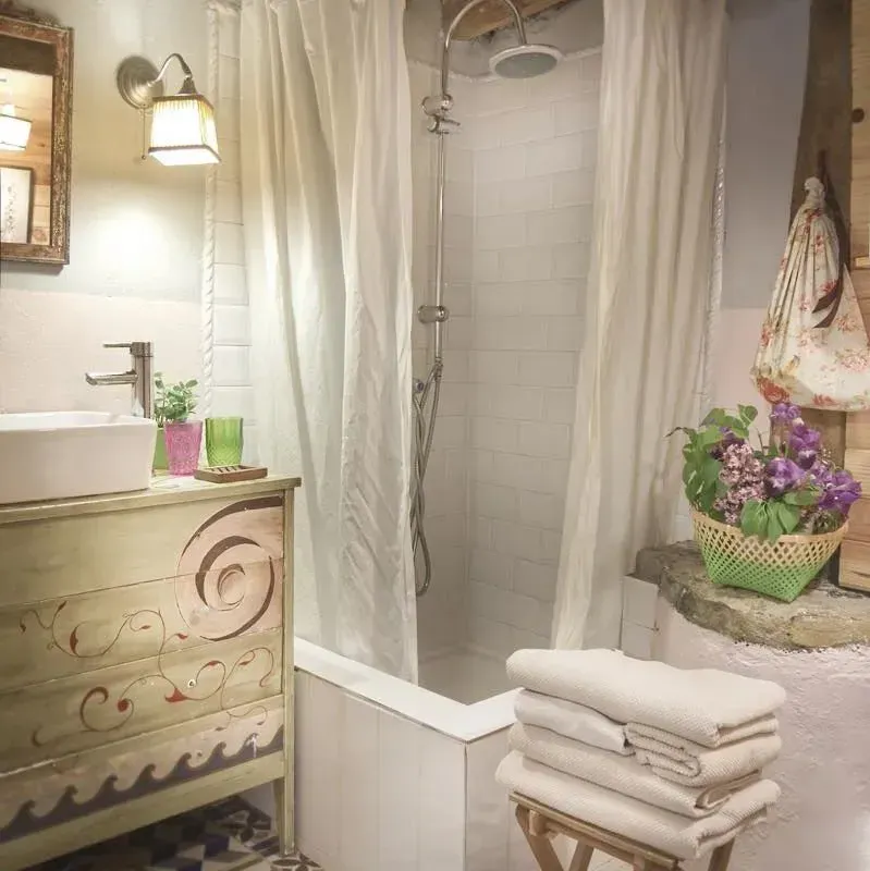 Baño con cortina de ducha blanca, lavabo blanco y un mueble decorativo. Toallas blancas apiladas sobre un taburete.